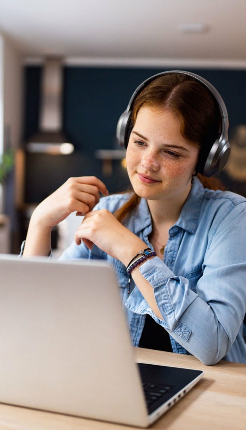 Teenage girl with headphones and laptop having online school class at home.